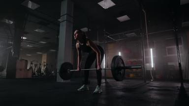 慢动作:一个女人在一个体重减轻的黑健身房里举起了一个杠铃.强壮的年轻女子举重锻炼身体